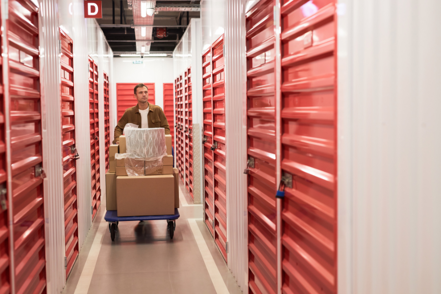 Man in jacket wheels dolly full of boxes through storage unit with orange doors. 
