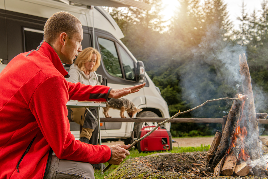 Man in red jacket sits with hand over fire next to woman with small dog beside her outside RV in the woods. 
