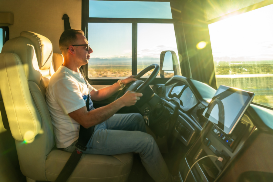 Man wearing jeans, t-shirt, and sunglasses drives RV at sunset with rays shining through vehicle. 