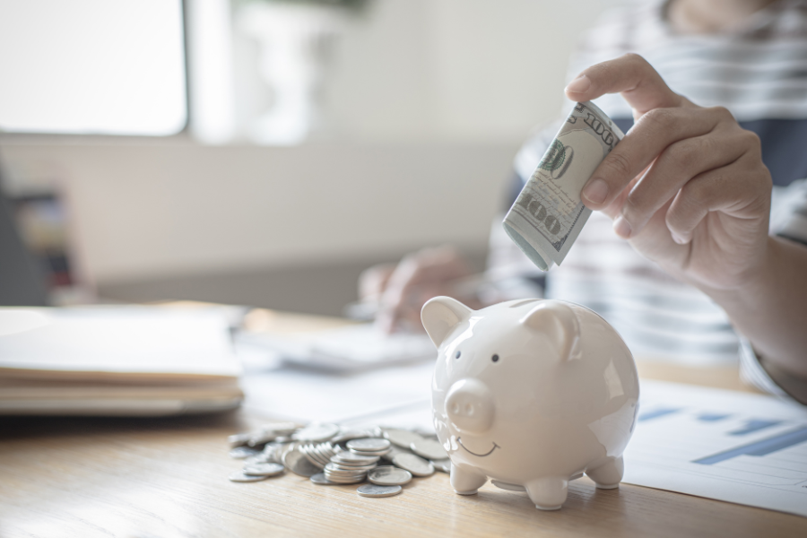 Woman in striped shirt puts folded 100 dollar bill in piggy bank sitting next to coin.
