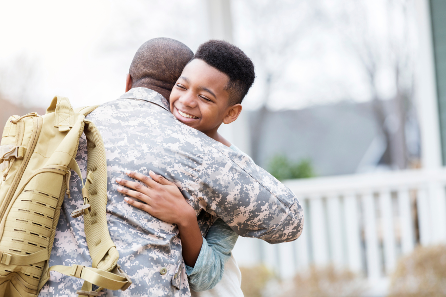 Boy smiles as he hugs his dad in military uniform with tactical backpack. 