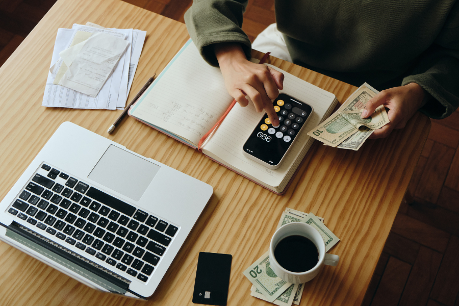 Aerial view of woman wearing green using phone as calculator while holding cash next to her computer, notebook, and coffee mug. 