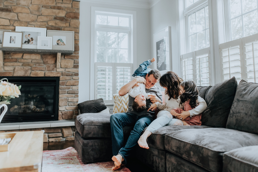 Family of four playing on dark gray couch with stone fireplace and lots of bright windows behind. 