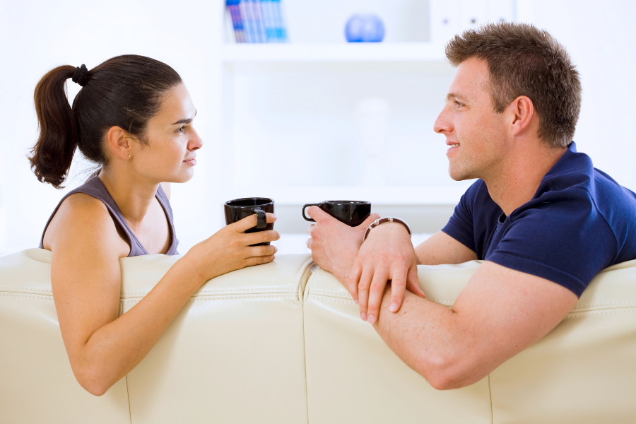 Man and woman sitting facing each other with cups of coffee talking on couch. 
