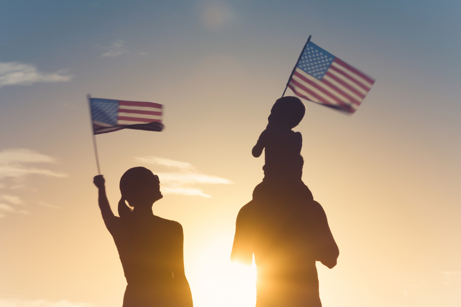 Silhouette of woman holding American flag in sunset sky stands next to man with child on shoulders holding American flag. 