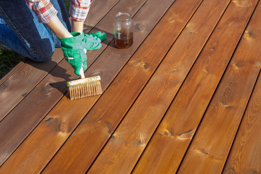 Person in jeans and plaid shirt wears green gloves and stains deck with brush. 