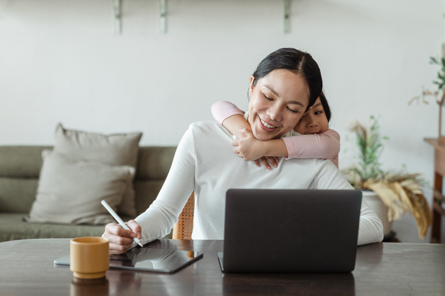 Woman in white long sleeve shirt sits with tablet and computer at table with little girl hanging around her neck. 
