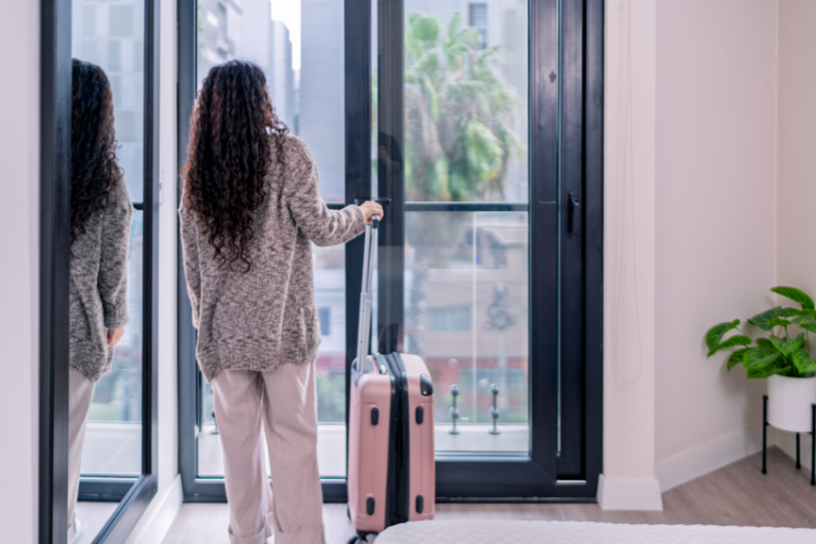 Woman wearing sweaters stands with her hand on her pick suitcase in room looking out of window to street beyond. 