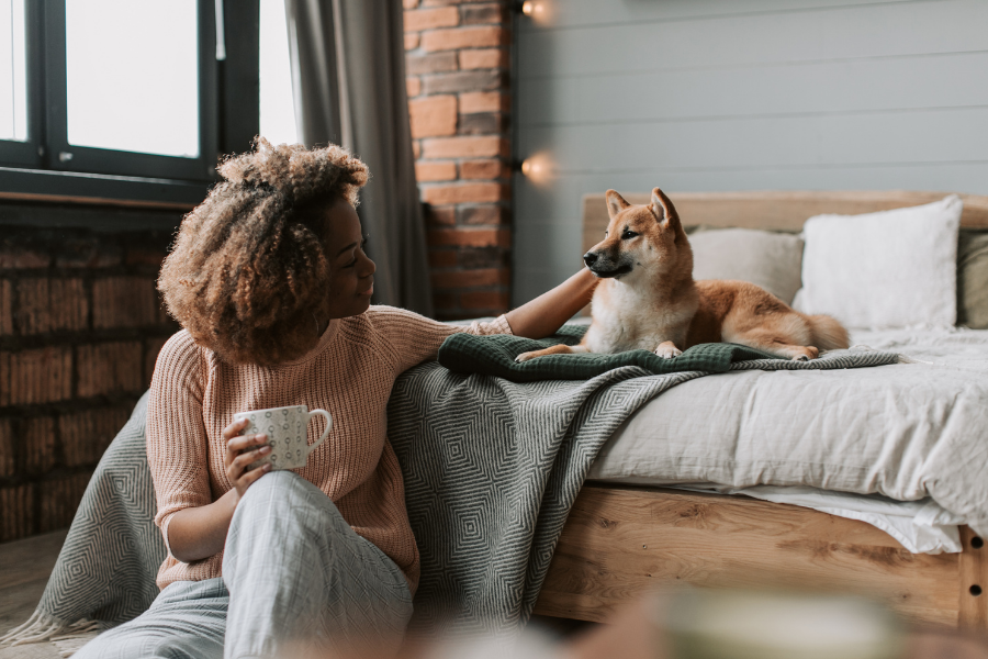 Woman sitting with mug on floor in front of bed pets dog laying on layered bedding. 