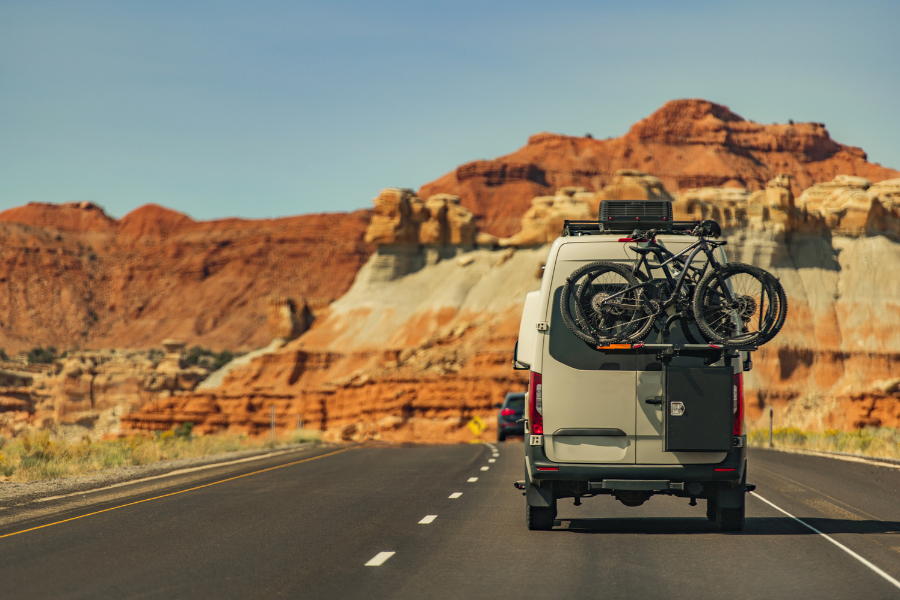 Rear view of sprinter camper can with bikes hitched to the back drive down two-lane highway in desert with red mountains. 