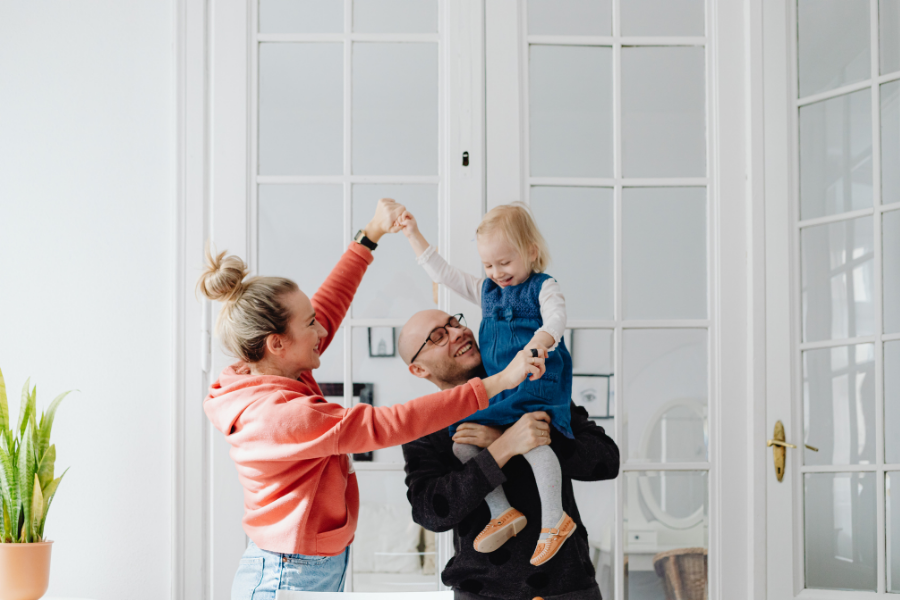 Mom playing with daughter in dad's arms in house with white walls and large windows.