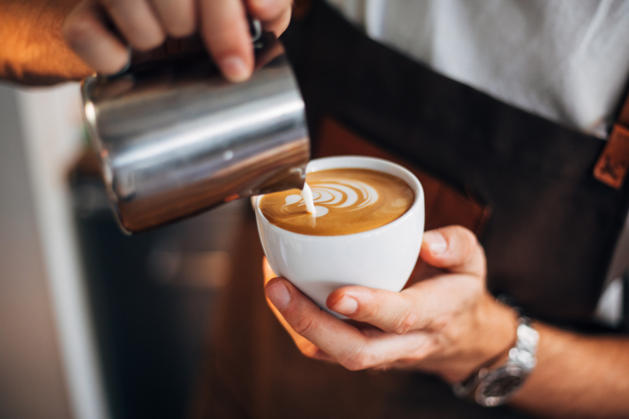 Male barista wearing silver watch and black apron pouring steamed milk into coffee to make latte art. 