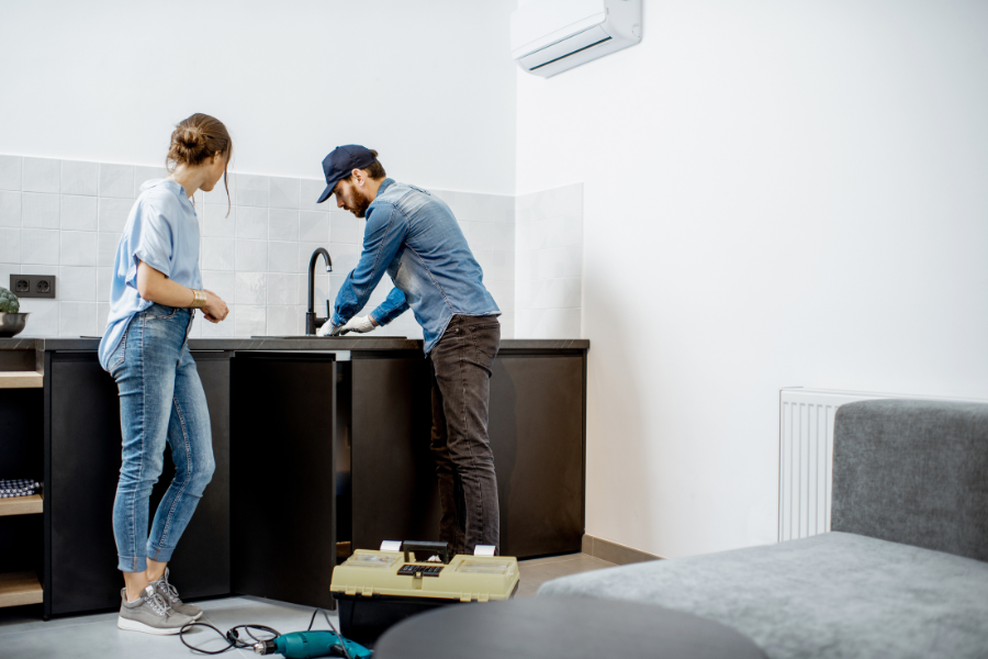 Woman in blue jeans watches man in denim shirt and ball cap fix sink. 