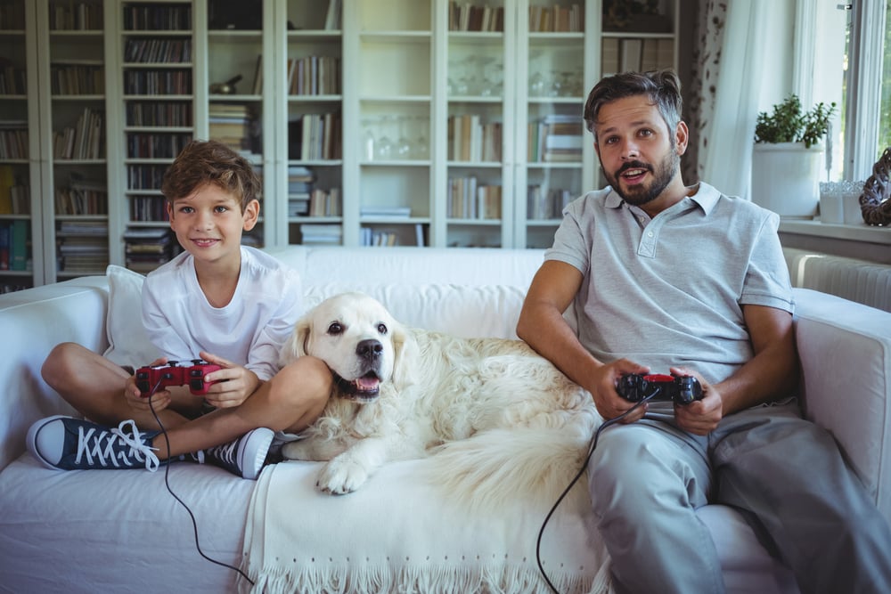Father and son sitting on sofa with pet dog and playing video games at rental home with their pet