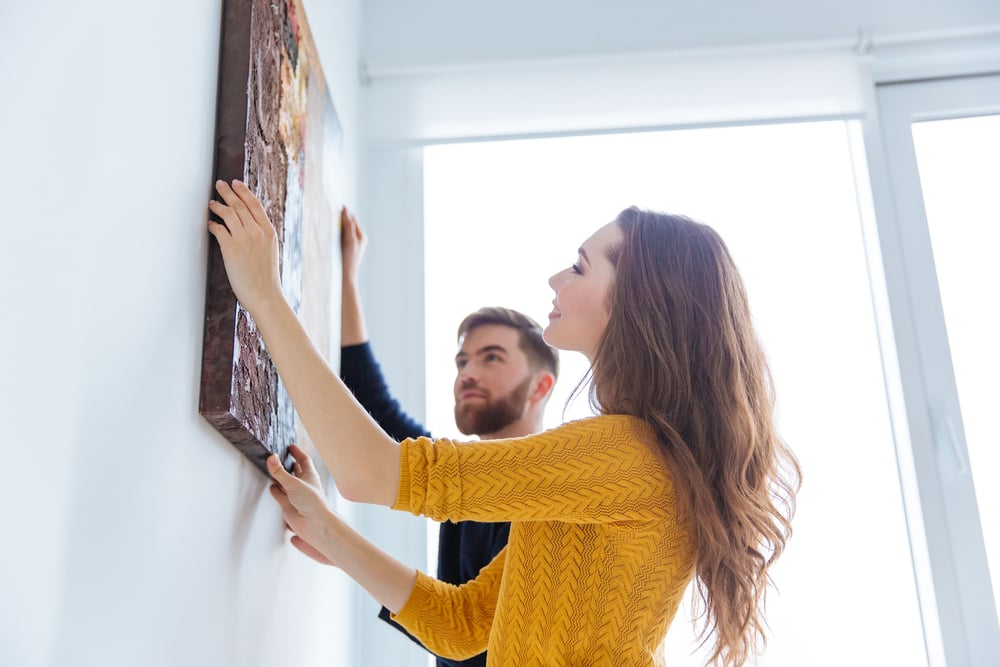 Woman wearing yellow sweater hangs art on the wall with man next to her. 