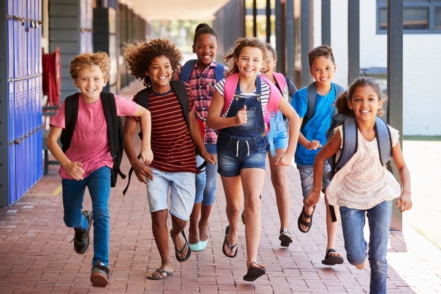 Group of diverse preteens wearing their backpacks smile as they run down brick path past blue school lockers.