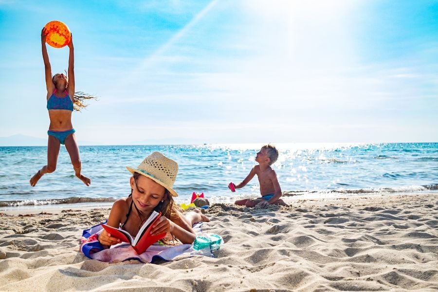 Three kids playing in bright sunshine at beach. One is jumping with an orange ball, one is shoveling sand while watching his sister, and the third is wearing a sun hat while laying down reading a book on her stomach. 