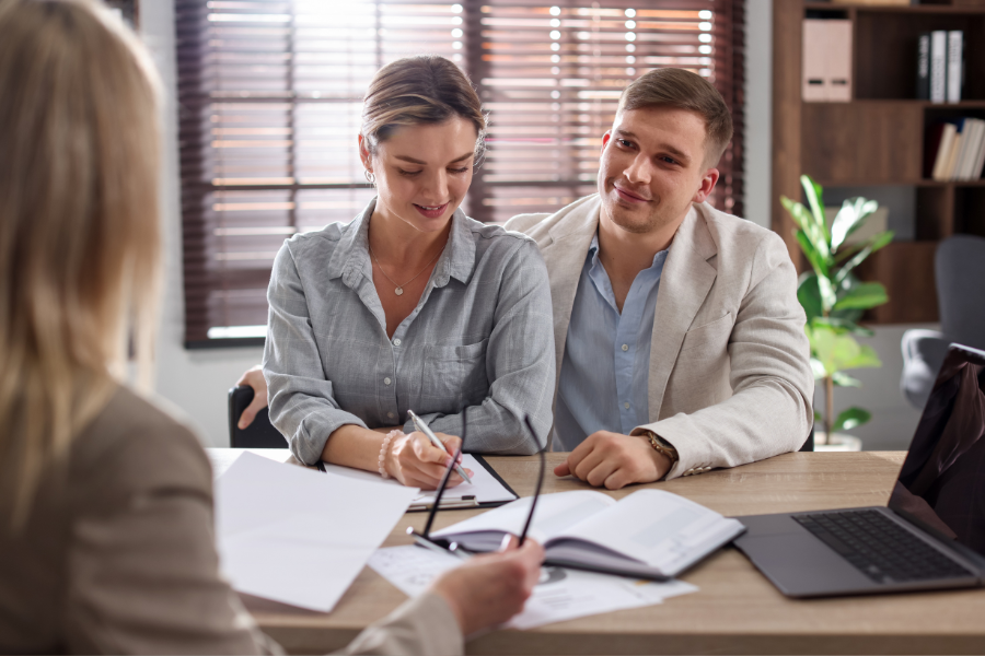 Woman in button down shirt smiles while signing paper sitting next to man in blazer smiling across the table at female professional holding glasses in hand.