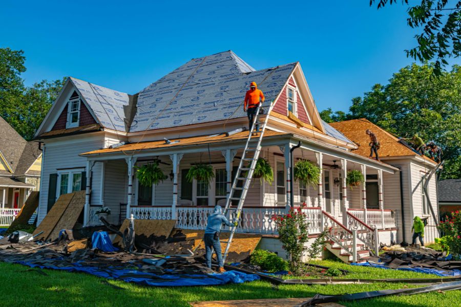 Man in orange shirt works roof of victorian home with man in blue hoodie standing at the base of the ladder. 