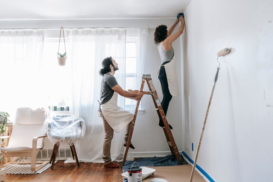 Man holding ladder steady while woman stands on it to place blue tape on wall to prep for paint.