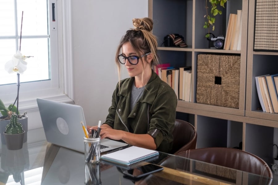 Woman in green jacket and black-rimmed glasses with hair in a bun, sits at glass desk typing on a computer. 