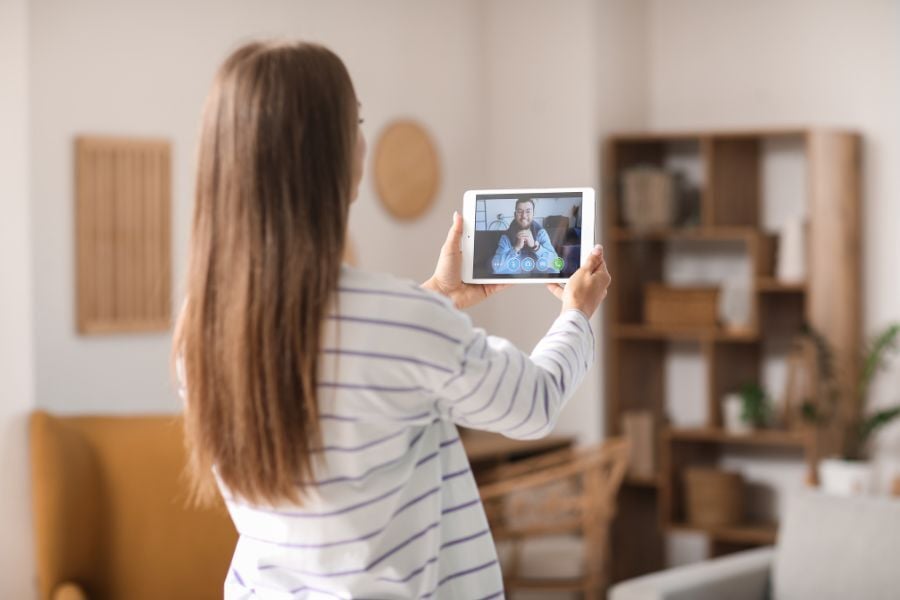 Woman in striped long sleeve shirt holds up tablet in room while video messaging. 