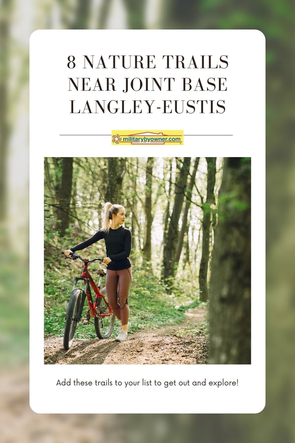 Woman with hair in a pony tail wearing a black long-sleeve shirt and brown leggings walks red mountain bike through trail in woods with text, 8 Nature Trails Near Joint Base Langley-Eustis