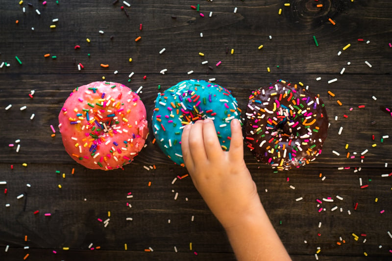 Photo of three sprinkles donuts on a wood table one pink, one blue, one chocolate with a hand reaching for the blue one in the middle. 