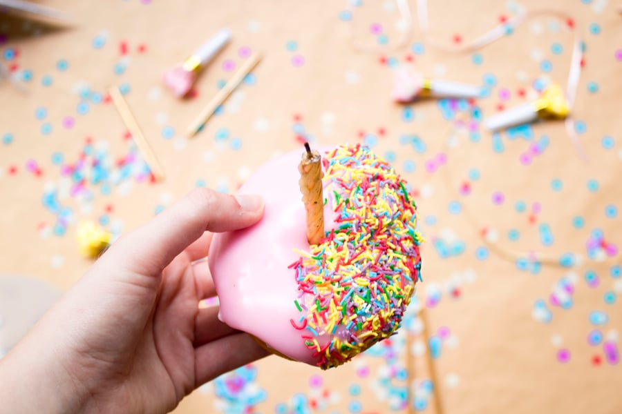 Photo of hand holding a pink iced donut with sprinkles on half and a gold candle in the center. 