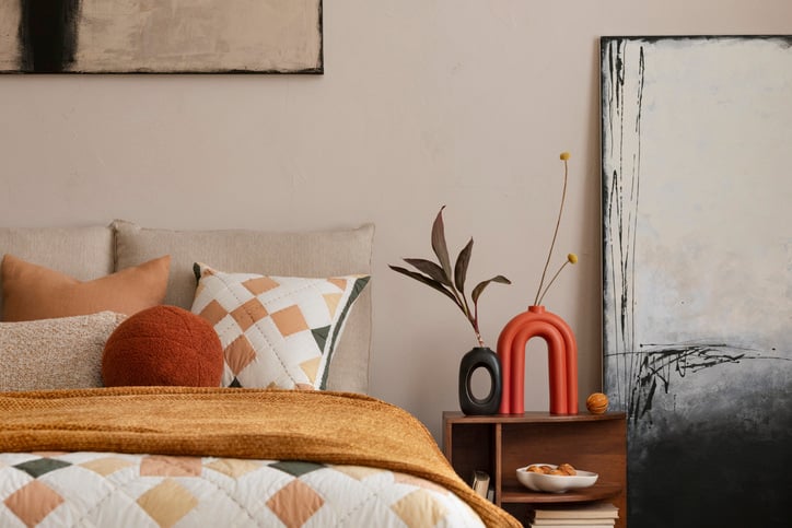 Stylish composition of orange bedroom interior with mock up poster frame, orange bedding, wooden bedside table, stylish sculpture, vase with leaves and personal accessories. Home decor.