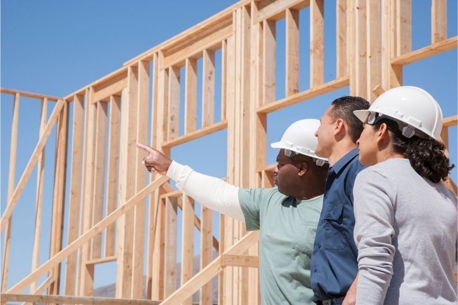 Man and woman in white hard hats point stand with man to look at new construction home. 