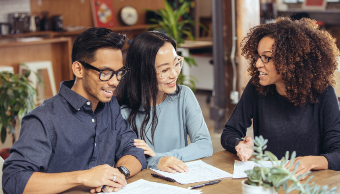 Man and woman wearing glasses sit at table with woman talking over paperwork. 