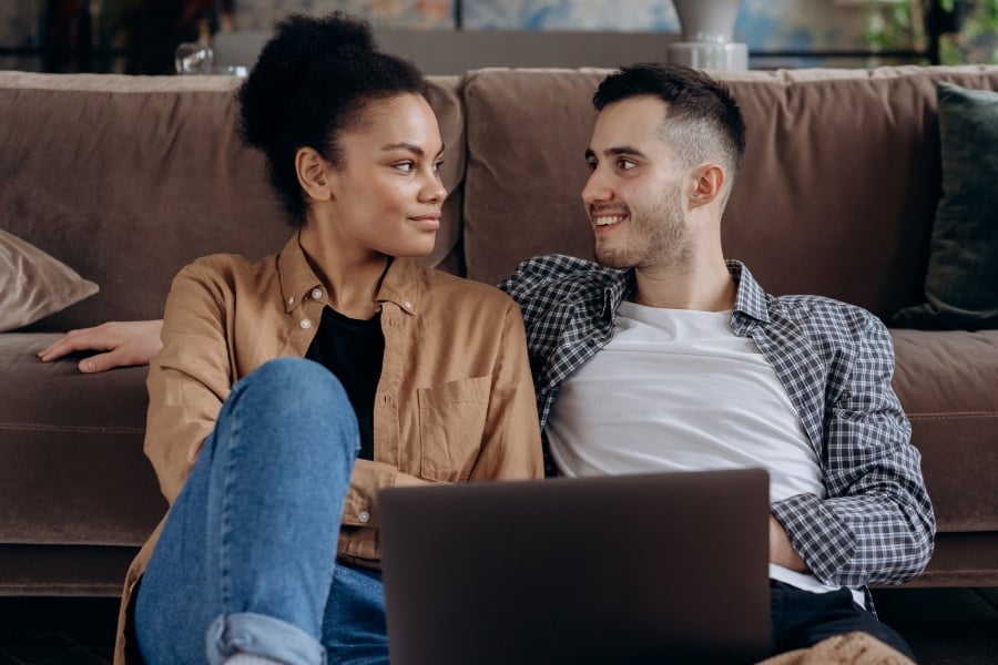 Man in white shirt and button down and woman with dark hair and brown shirt look at each other while holding a computer on their laps. 