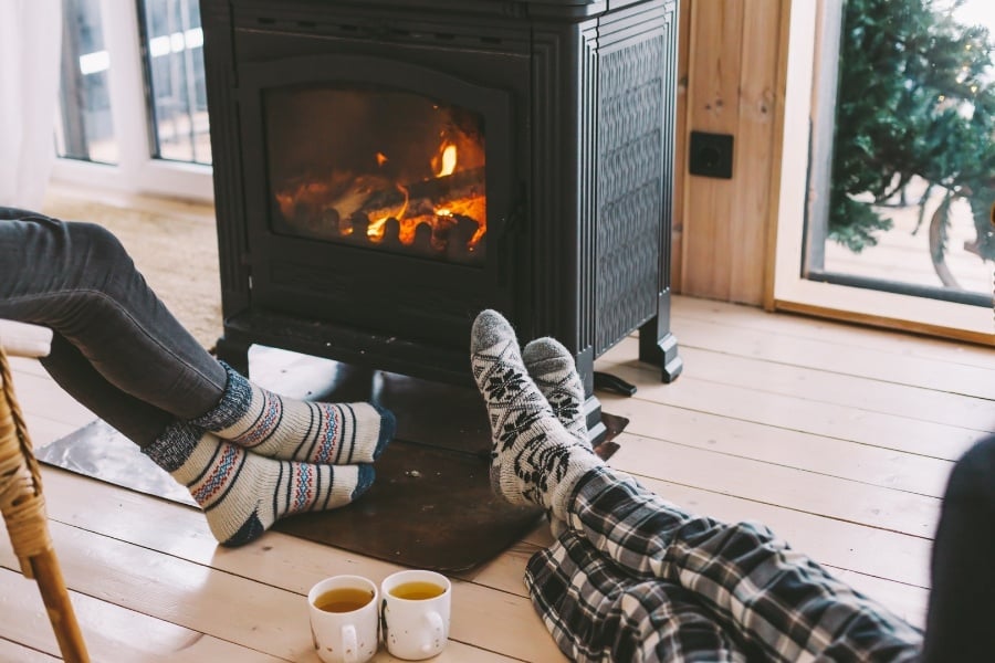 Two pairs of legs and feet with striped and plaid relaxing next to burning black fireplace with two filled white mugs between them. 