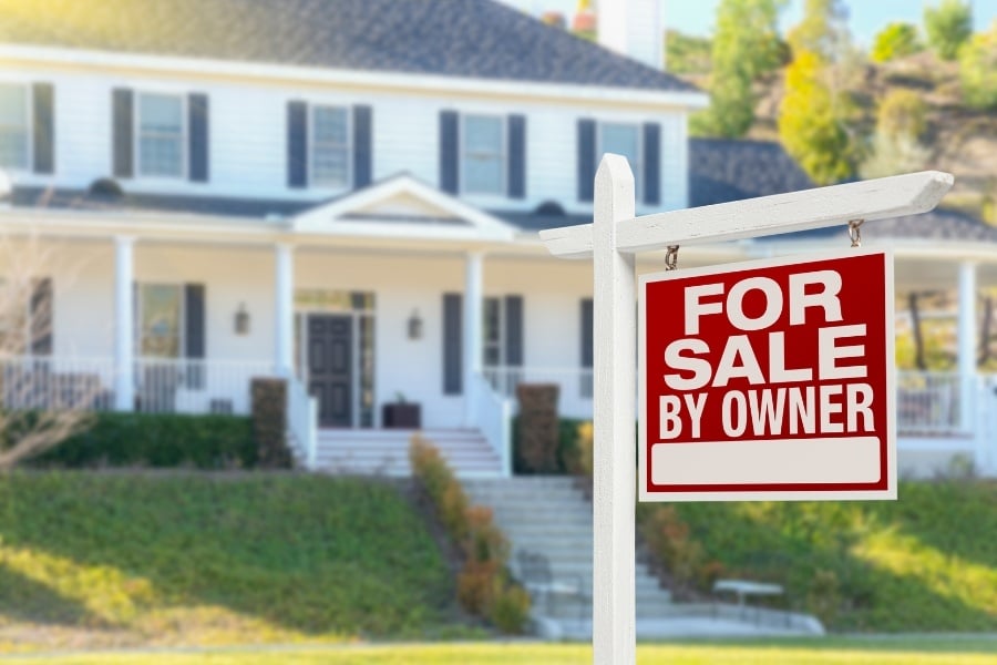 Photo of out of focus large white home with lots of windows and a For Sale By Owner sign hanging from white post in the front yard. 