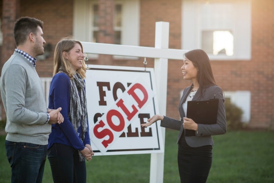 Man and woman in blue shirt and scarf stand with woman real estate agent in front of home with for sale sign marker sold in red letters