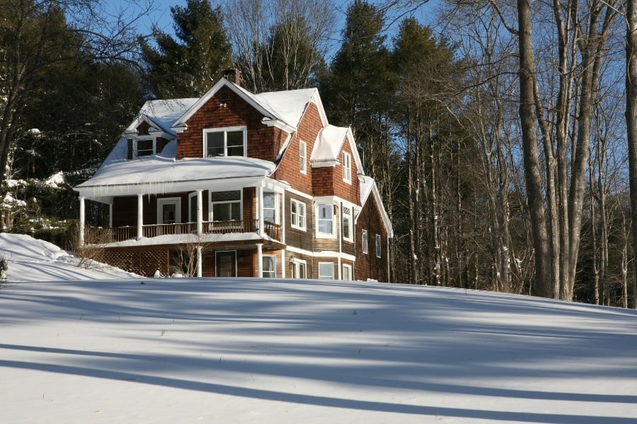 Wooden house with white trim, snow on the ground, trees in the background and sun shining.