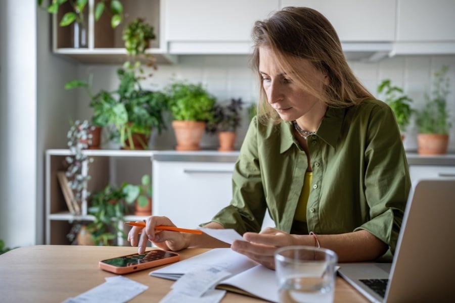 Woman in green shirt looking at papers and typing calculator on phone. 