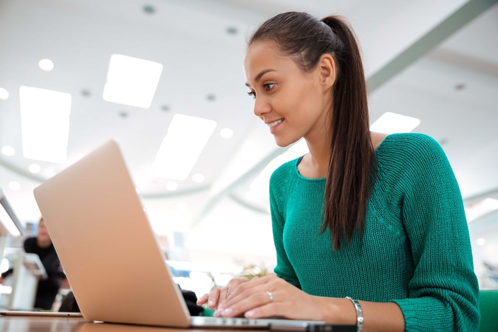 Woman with long ponytail and green shirt smiles at computer on desk.