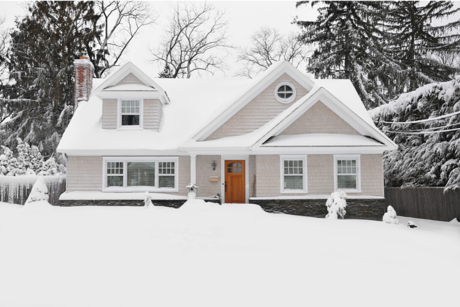 Light-colored house with bright orange door covered in snow with a blanket of snow on the ground and tall trees in the back. 