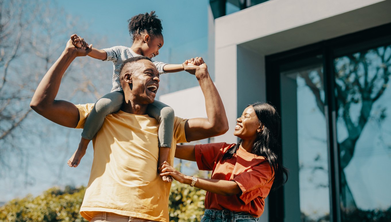 Dad carrying his daughter on his shoulders and standing next to mom outside home